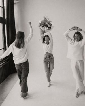 Three women in a room with one holding flowers, all wearing white tops.